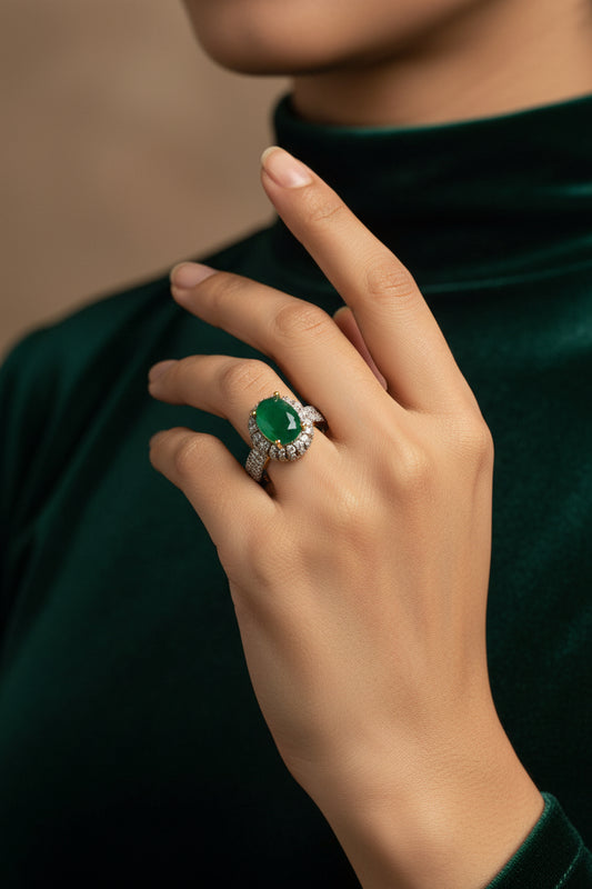 A yellow gold ring with a green oval gemstone and pavé-set clear stones, displayed on a white textured surface inside a decorative frame. A white size tag is attached to the ring.