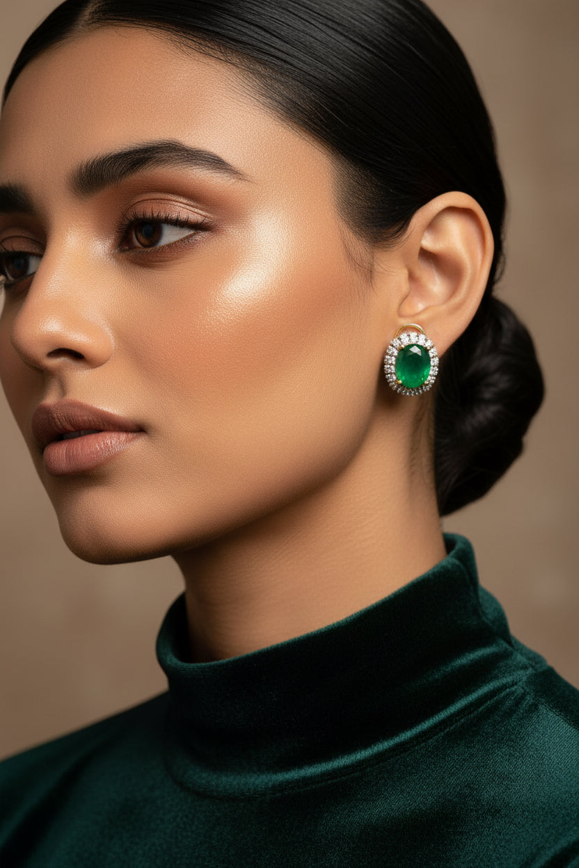 A pair of gold hoop earrings with oval green gemstones surrounded by clear stones, displayed on a white textured surface inside a decorative frame.