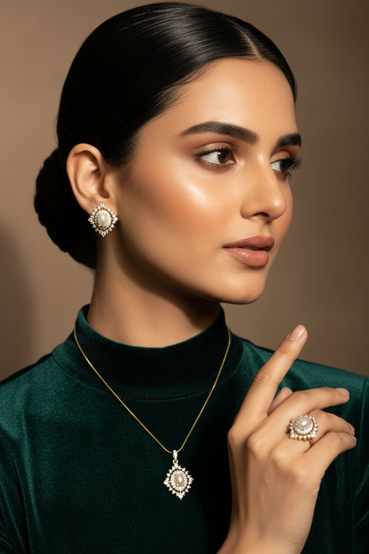 A jewelry set including a pendant necklace, ring, and earrings, displayed on a hand wearing a black glove. The jewelry features white pearls surrounded by gold and clear crystals, set against a neutral light background.