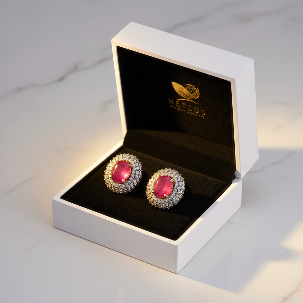 A pair of gold-plated stud earrings featuring a heart-shaped ruby center stone surrounded by diamond cut patterns, displayed on a beige cushion with the brand Metcos Designer Jewelry visible.