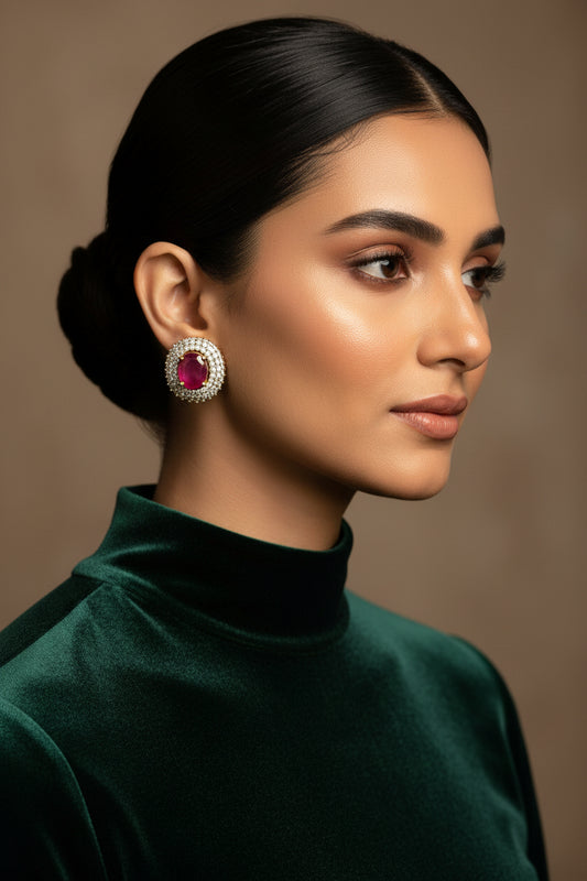 A pair of gold-plated stud earrings featuring a heart-shaped ruby center stone surrounded by diamond cut patterns, displayed on a beige cushion with the brand Metcos Designer Jewelry visible.