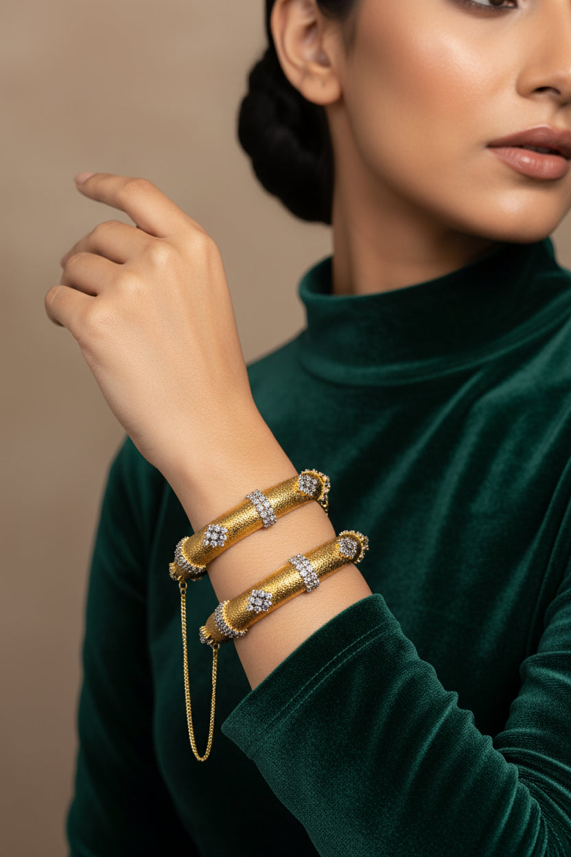 Pair of 24K gold plated kara bangles with a textured diamond-cut body, three American zircon bands and a safety chain, worn on the wrist